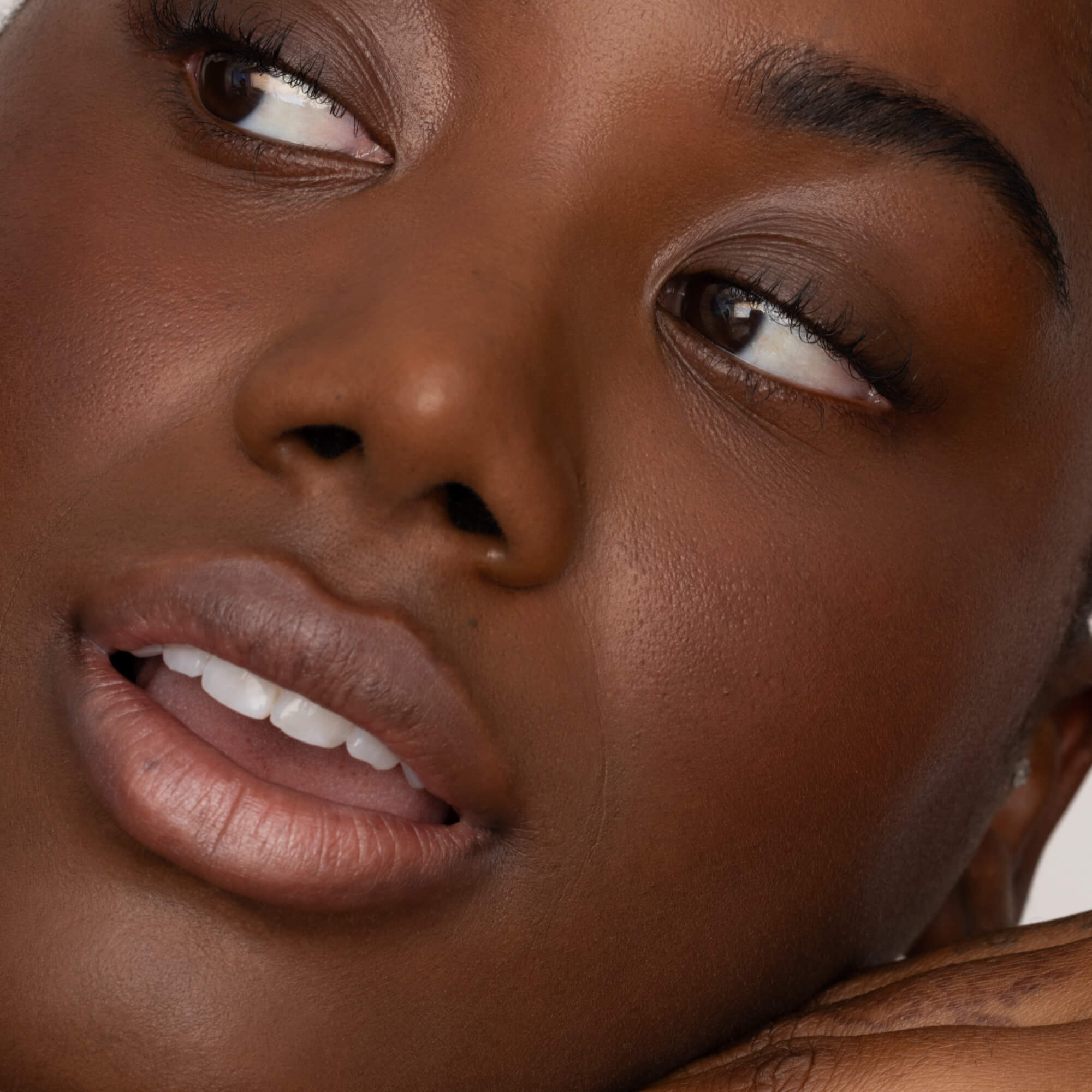 A close-up of a woman’s glowing brown skin, natural makeup, and soft pink lips from Faire’s Poppy & Pout Lip Tint in Farrah. She looks off to the side with a gentle, relaxed expression.