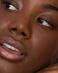A close-up of a woman’s glowing brown skin, natural makeup, and soft pink lips from Faire’s Poppy & Pout Lip Tint in Farrah. She looks off to the side with a gentle, relaxed expression.