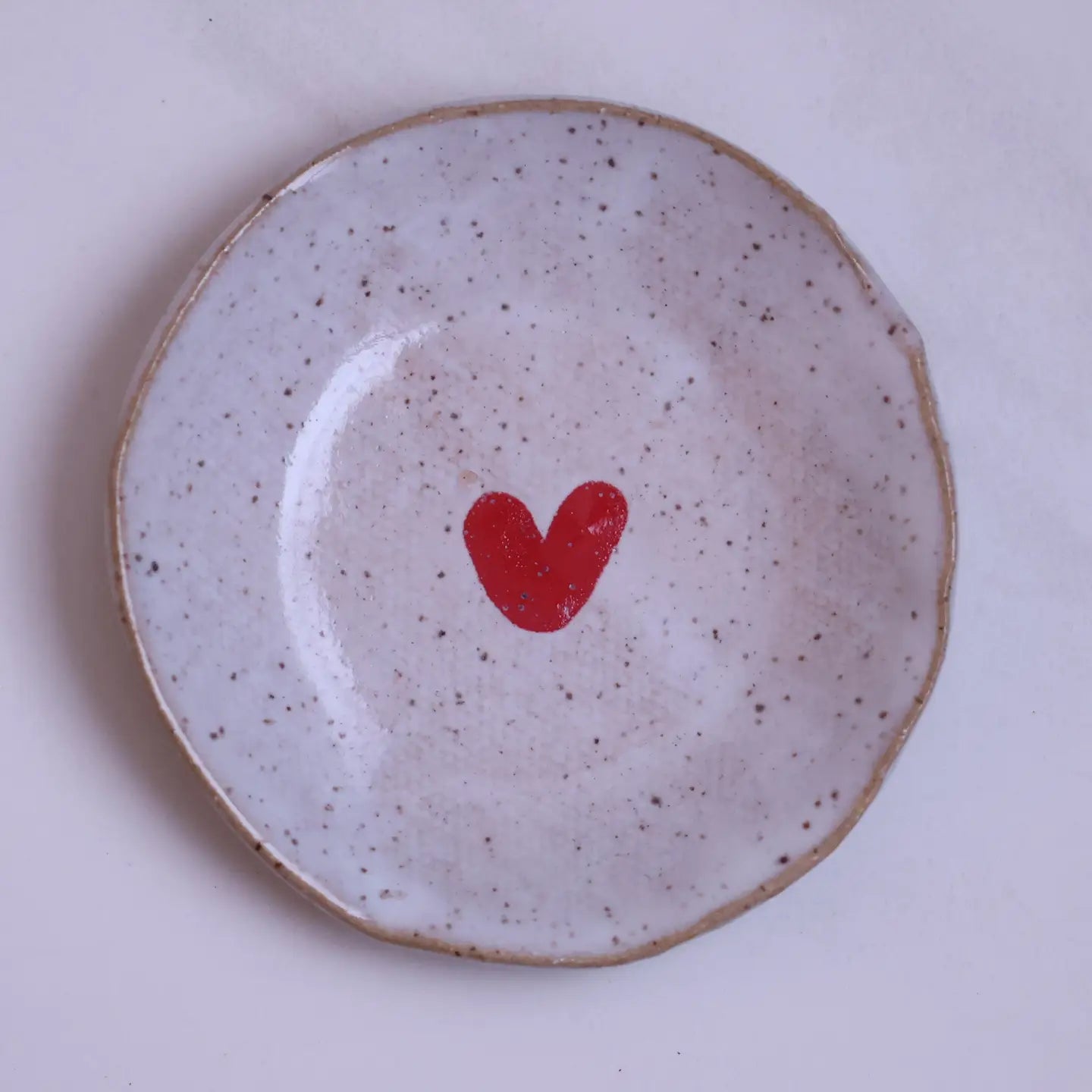 The Faire Mini Plate is a round, speckled ceramic dish with a small red heart in the center, ideal for holding jewelry or snacks. Photographed on a plain light background.