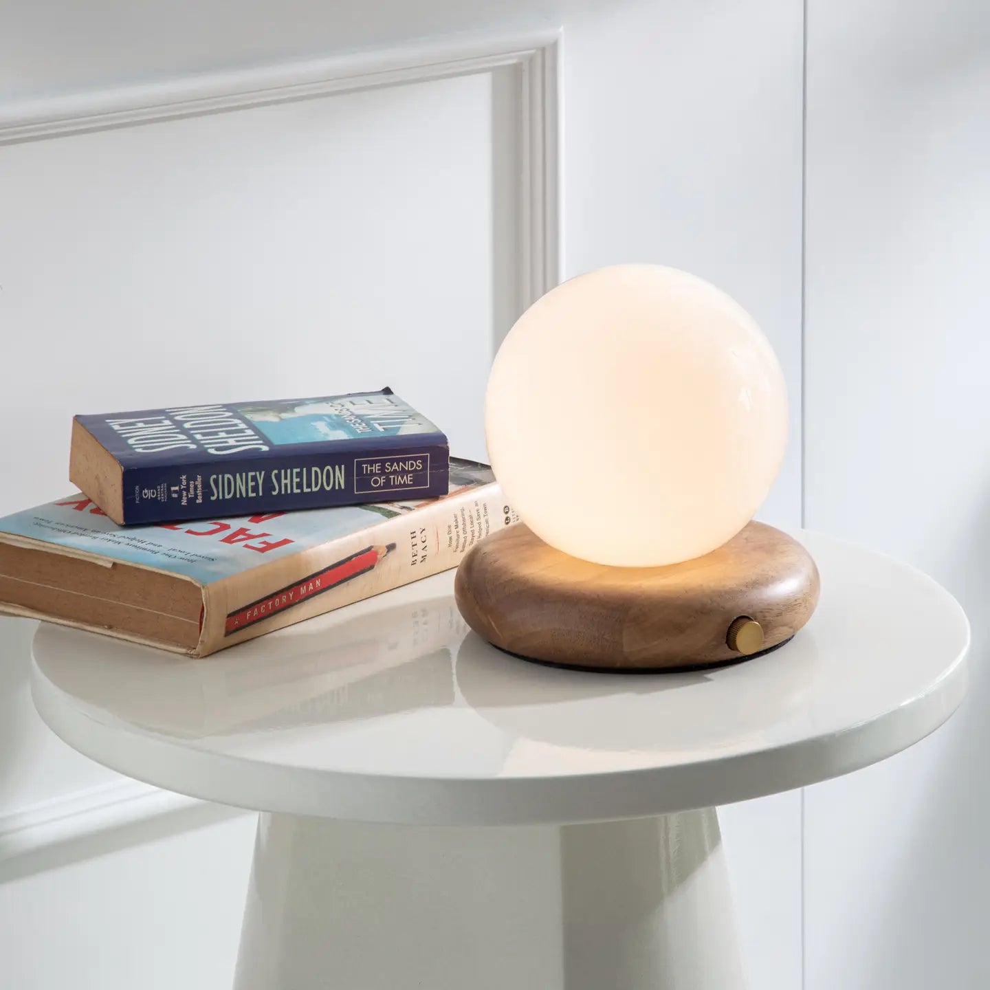 The Faire Valencia Portable Light with a wooden base rests on a small white table beside two stacked books, including one by Sidney Sheldon, creating a bright and minimalistic scene.