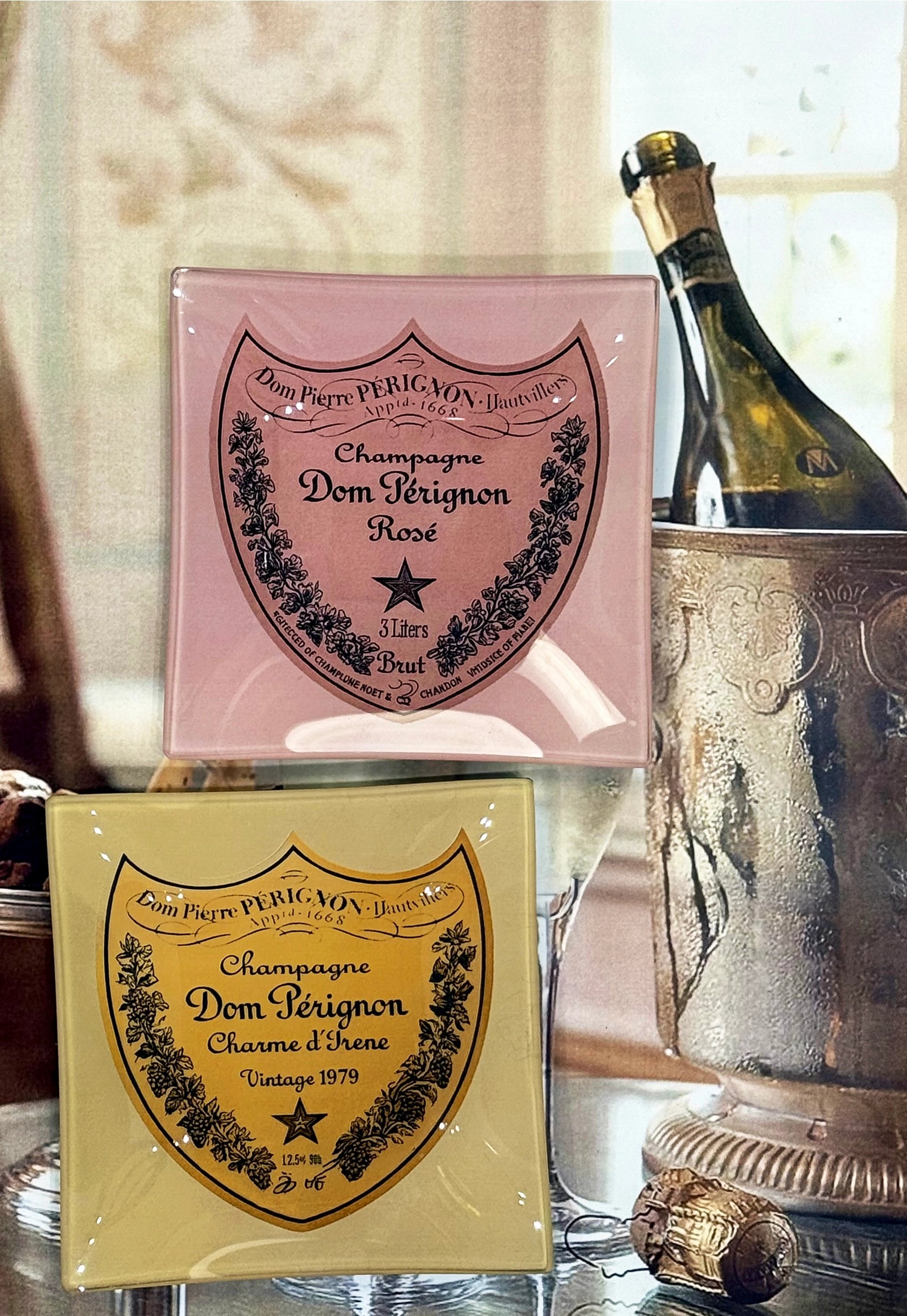 Two square Faire Dom Perignon Brut Glass Trays, one pink for Rosé and one gold for Vintage 1979, are displayed before a champagne bottle and ice bucket—each a handcrafted piece inspired by the iconic labels.