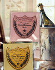 Two square Faire Dom Perignon Brut Glass Trays, one pink for Rosé and one gold for Vintage 1979, are displayed before a champagne bottle and ice bucket—each a handcrafted piece inspired by the iconic labels.