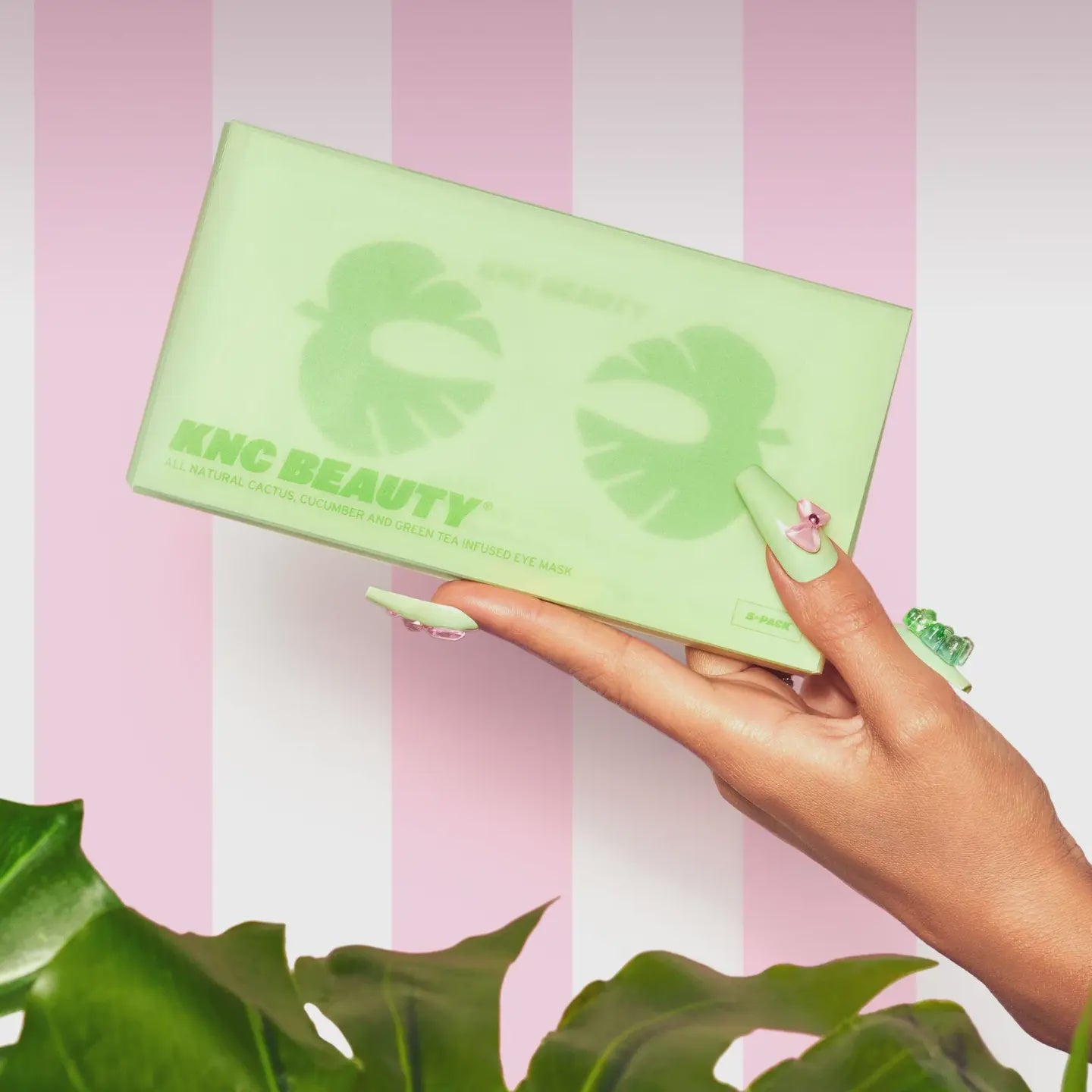 A manicured hand displays a green Faire Cucumber/Tea Eye Mask package against a pink and white striped backdrop, emphasizing its natural ingredients. Green leaves are partly visible at the bottom.