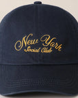 The Faire New York Social Club Baseball Cap in navy blue has "New York Social Club" embroidered in yellow cursive on the front and features an adjustable strap.