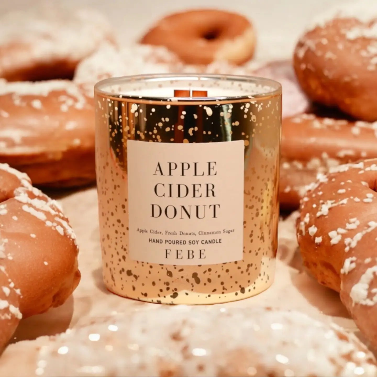 A gold FEBE Candle by Faire, labeled "Apple Cider Donut," is nestled among frosted donuts, with its jar adorned in silver speckles. Its subtle smokey fragrance evokes a warm, autumnal ambiance.