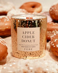 A gold FEBE Candle by Faire, labeled "Apple Cider Donut," is nestled among frosted donuts, with its jar adorned in silver speckles. Its subtle smokey fragrance evokes a warm, autumnal ambiance.