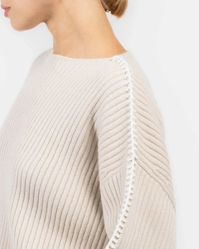 A person wears the Rag & Bone ingrid boat neck sweater in light beige, featuring chunky ribbed knit and white stitching along the sleeve seam, shown from the side with their face partially out of frame.