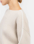 A person wears the Rag & Bone ingrid boat neck sweater in light beige, featuring chunky ribbed knit and white stitching along the sleeve seam, shown from the side with their face partially out of frame.