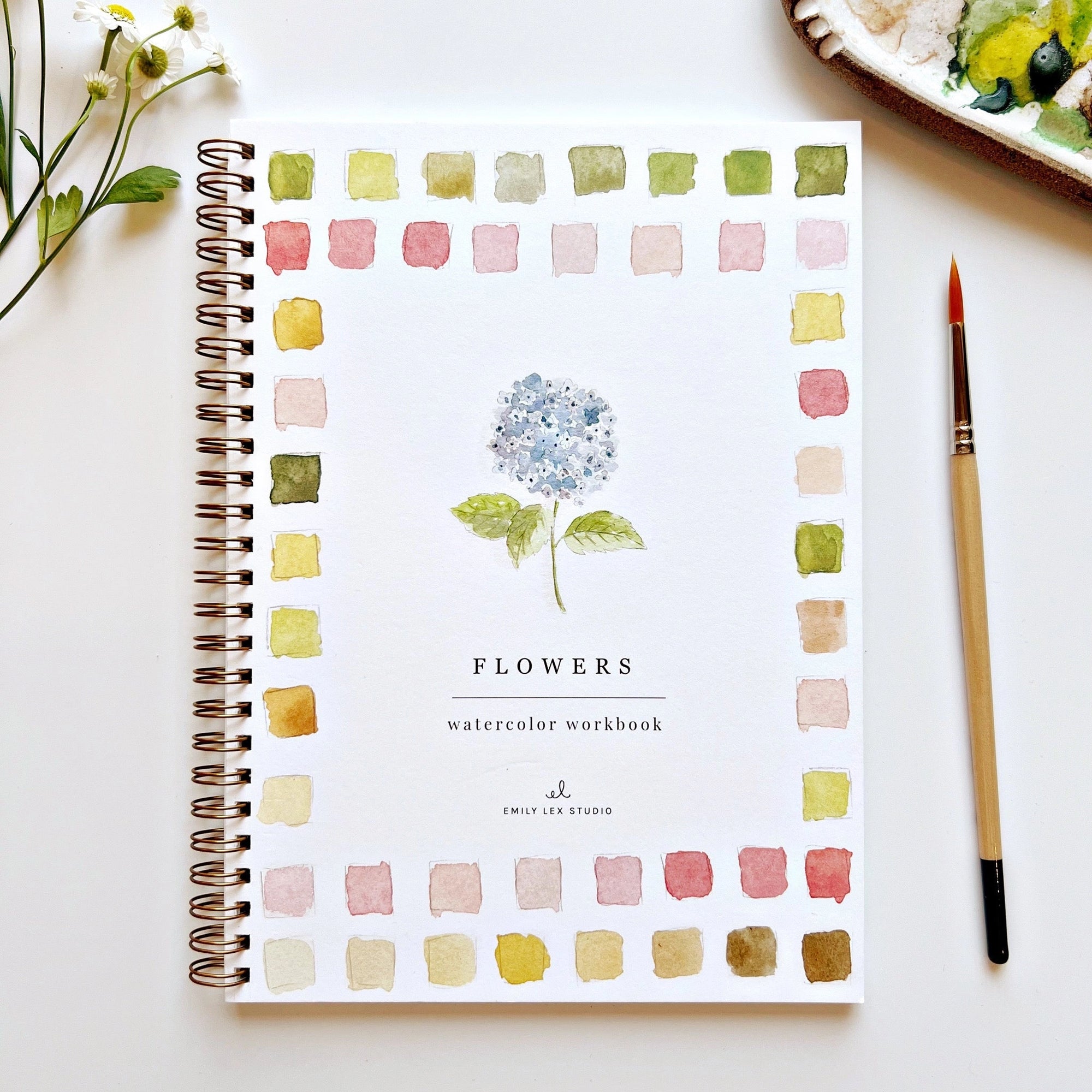 The Faire Watercolor Workbook, spiral-bound and beginner-friendly, features a blue hydrangea cover with colorful borders and sits near a paintbrush, palette, and white flowers—perfect for aspiring artists.