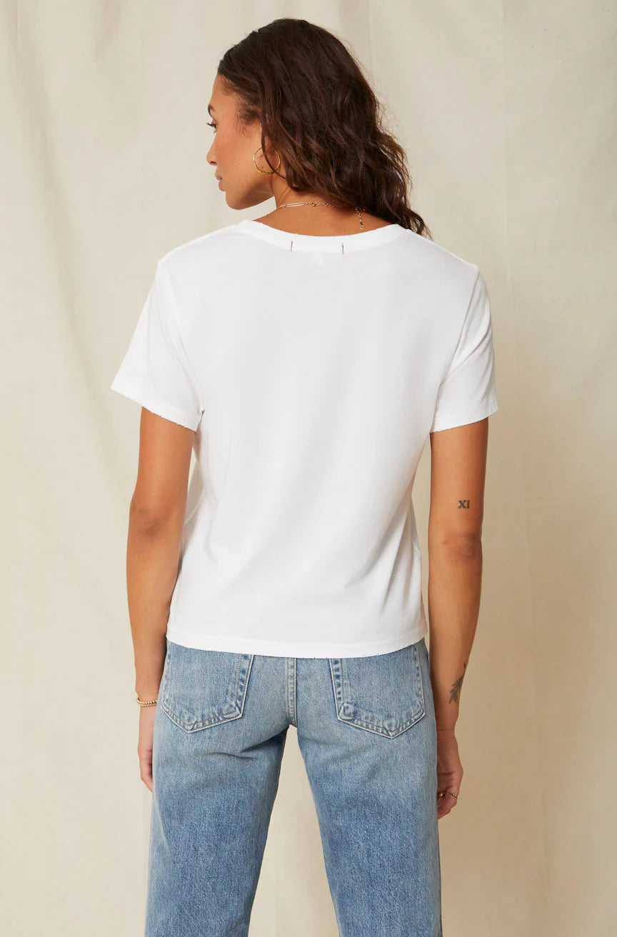 A person with long wavy hair, a small left arm tattoo, and blue jeans faces away from the camera wearing AMO Denim's Classic Tee, featuring distressed edges. The 100% cotton jersey shirt contrasts gently against a light-colored curtain in the background.