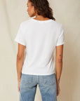 A person with long wavy hair, a small left arm tattoo, and blue jeans faces away from the camera wearing AMO Denim's Classic Tee, featuring distressed edges. The 100% cotton jersey shirt contrasts gently against a light-colored curtain in the background.