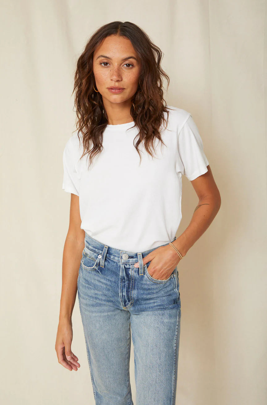 A person with long, wavy hair stands against a neutral background, wearing an AMO Denim Classic Tee made from 100% cotton jersey and blue jeans, their hands relaxed by their sides.