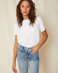 A person with long, wavy hair stands against a neutral background, wearing an AMO Denim Classic Tee made from 100% cotton jersey and blue jeans, their hands relaxed by their sides.