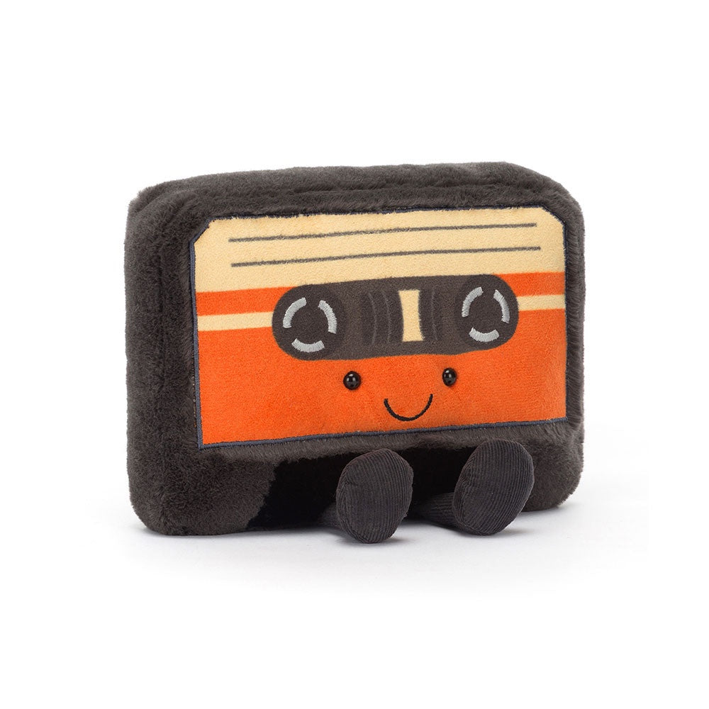 The Amuseables Cassette Tape by Jelly Cat Inc. is a plush toy shaped like a retro cassette tape. It includes fun details such as a smiling face and tiny black legs, along with its distinctive black and orange design featuring a beige striped label area. Constructed from soft polyester fibers, it makes for the perfect nostalgic cuddle companion.