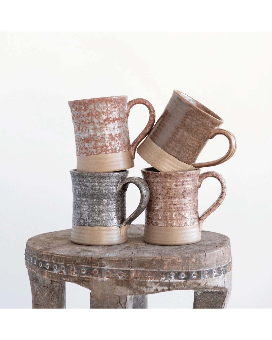 Four Creative Co-op Stoneware Mugs, each 16 oz. and showcasing earthy tones and rustic textures, are stacked on an aged wooden stool. Variations in brown, gray, and red enhanced by a reactive glaze stand out against a plain white backdrop.