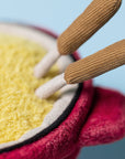 Close-up of the Amuseables Cheese Fondue by Jelly Cat Inc.—a plush toy shaped like a red fondue pot with a fuzzy yellow interior and brown handles, perfect for Switzerland fans or cozy dinner party décor.
