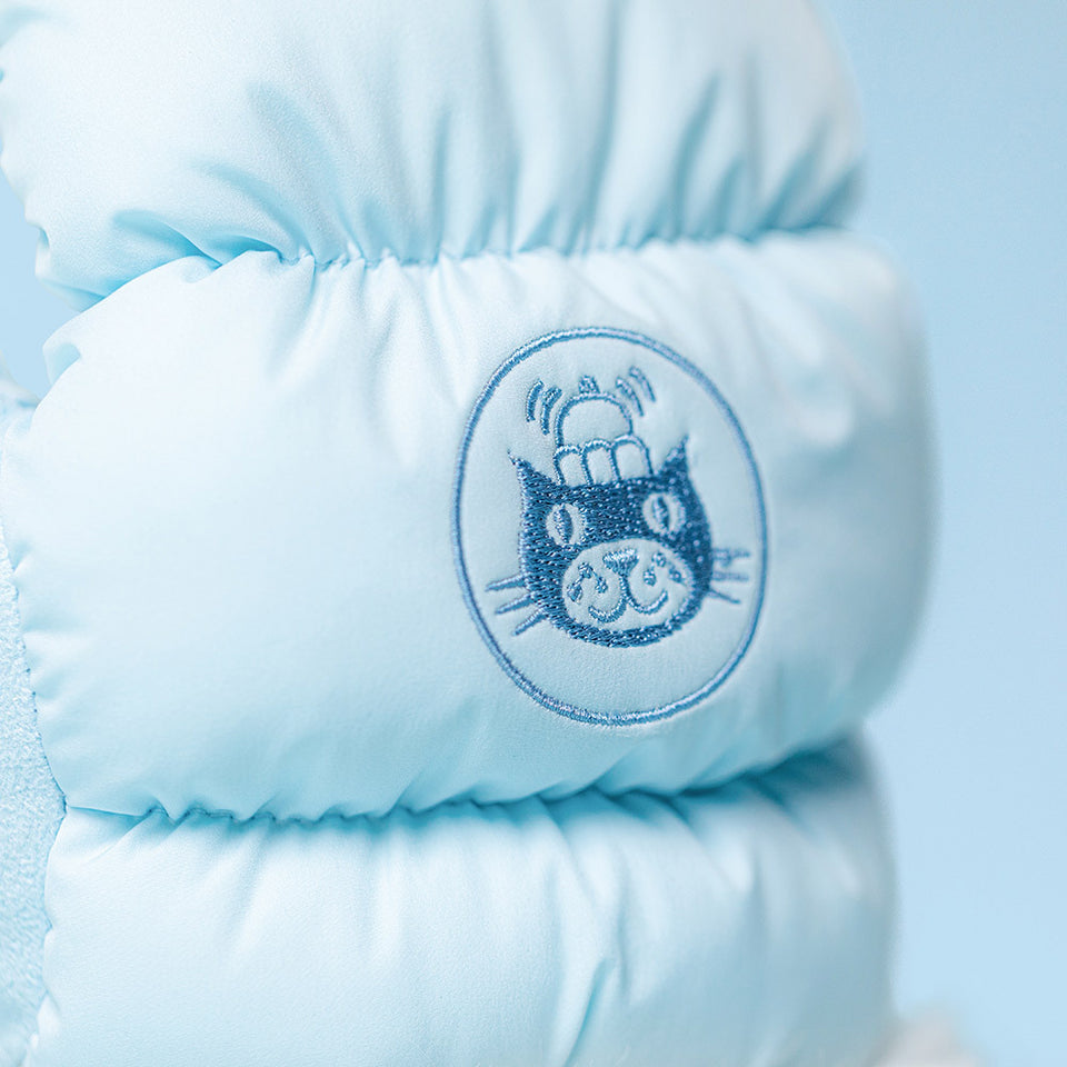 Close-up of the Jelly Cat Inc. Amuseables Mitten light blue puffy jacket featuring an embroidered cartoon cat face with stacked hats and a mitten, set in a circle on a soft blue background.