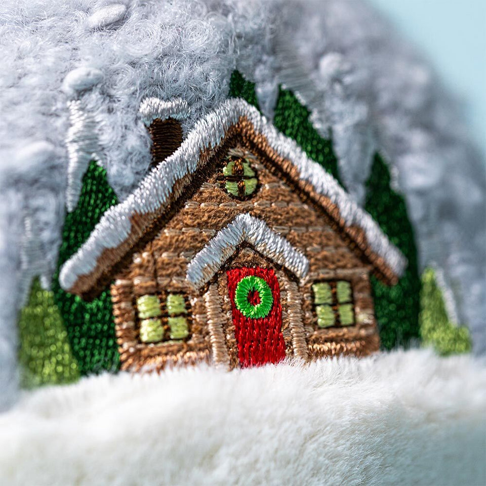 Close-up of Jelly Cat Inc.'s Amuseables Snow Globe: an embroidered log cabin with a snowy roof, green trees, and a red door with a wreath on soft white and gray fabric—a cozy ski-themed gift for après ski fans.