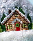 Close-up of Jelly Cat Inc.'s Amuseables Snow Globe: an embroidered log cabin with a snowy roof, green trees, and a red door with a wreath on soft white and gray fabric—a cozy ski-themed gift for après ski fans.