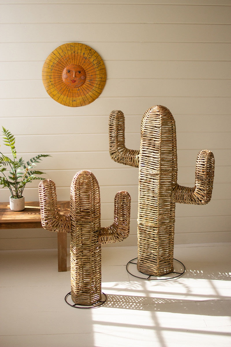 Three handmade seagrass cactus sculptures in varying sizes stand in a sunlit room of a Scottsdale, Arizona bungalow with a white wall, beside a wooden table with a small plant, under a circular wicker artwork featuring a sun design. (Kalalou, Inc)