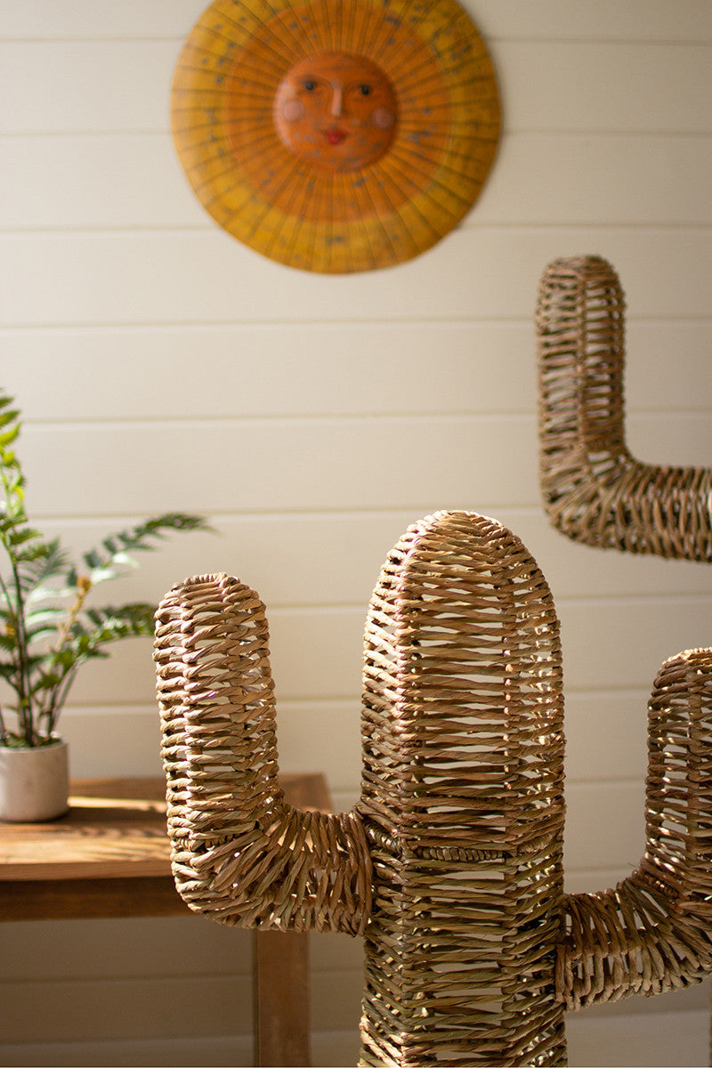 A Handmade Seagrass Cactus from Kalalou, Inc stands in the foreground of a warmly lit bungalow, with a sun decoration on the wall and a small plant on a wooden bench in the background.