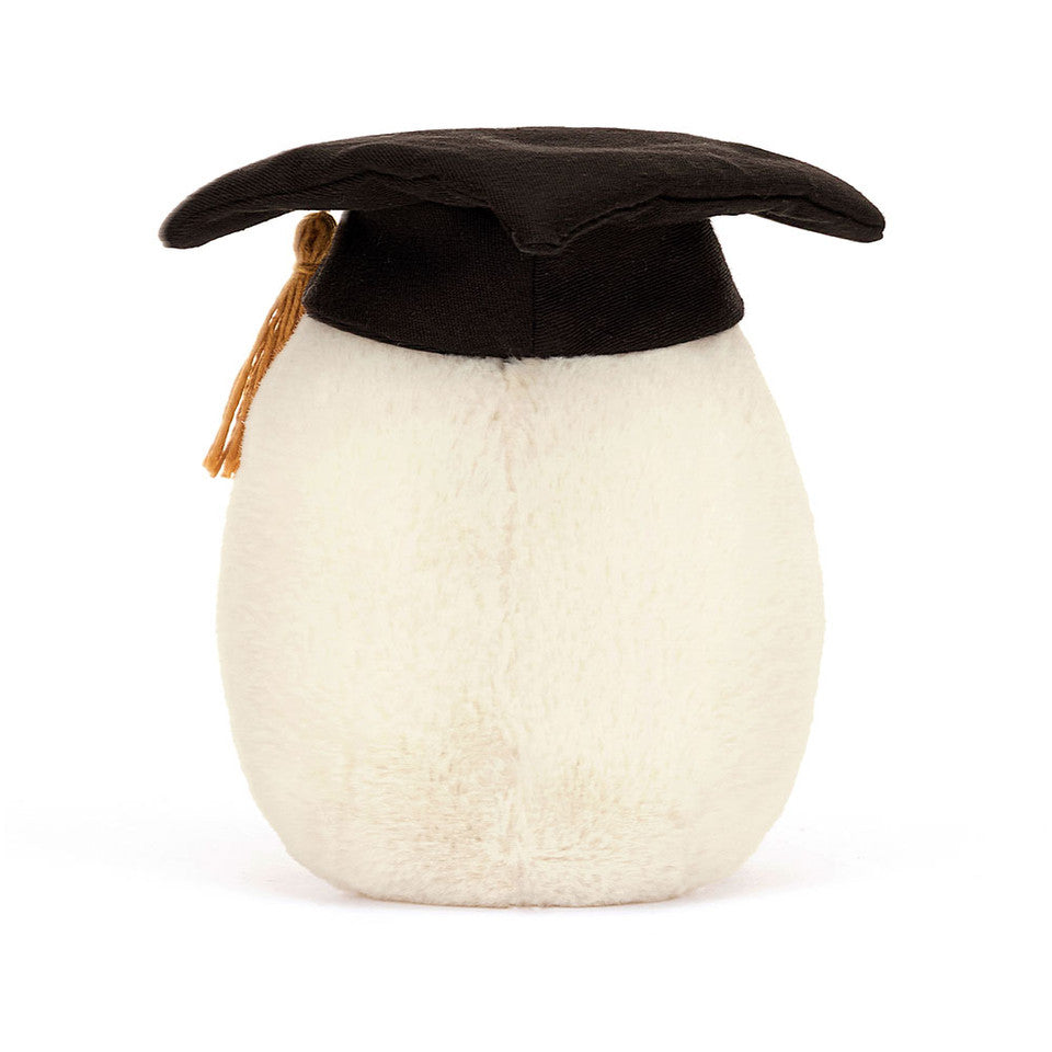 The Amuseables Boiled Egg Graduation by Jelly Cat Inc. is a plush toy shaped like a boiled egg, featuring soft fur and wearing a black cotton mortarboard with a gold tassel, set against a plain white background.