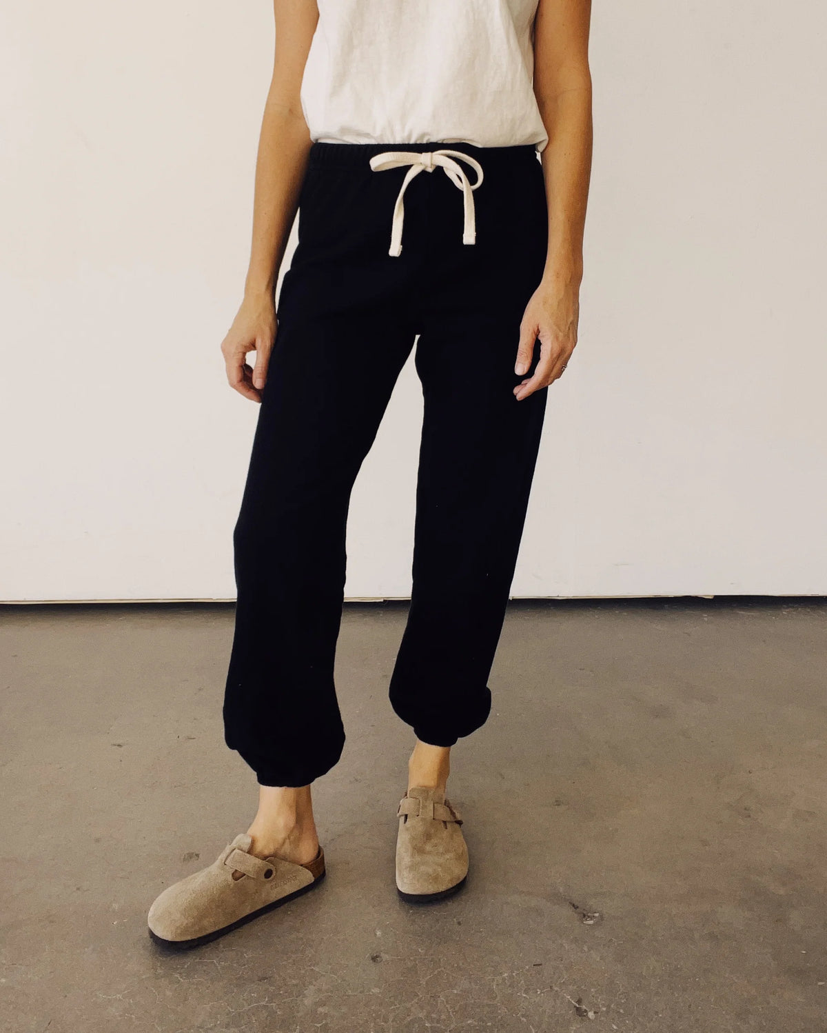 Hey Gang black sweatpants styled casually with a white top and tan slip-ons on a concrete surface.