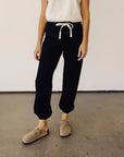 Hey Gang black sweatpants styled casually with a white top and tan slip-ons on a concrete surface.