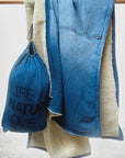 The Free City (sparrow, LLC) Indigo Sherpa Blanket/Indigo Blues, featuring soft white sherpa lining, is draped over a rod beside a blue "LIFE NATURE LOVE" bag. Part of the art commerce logo appears at the bottom.