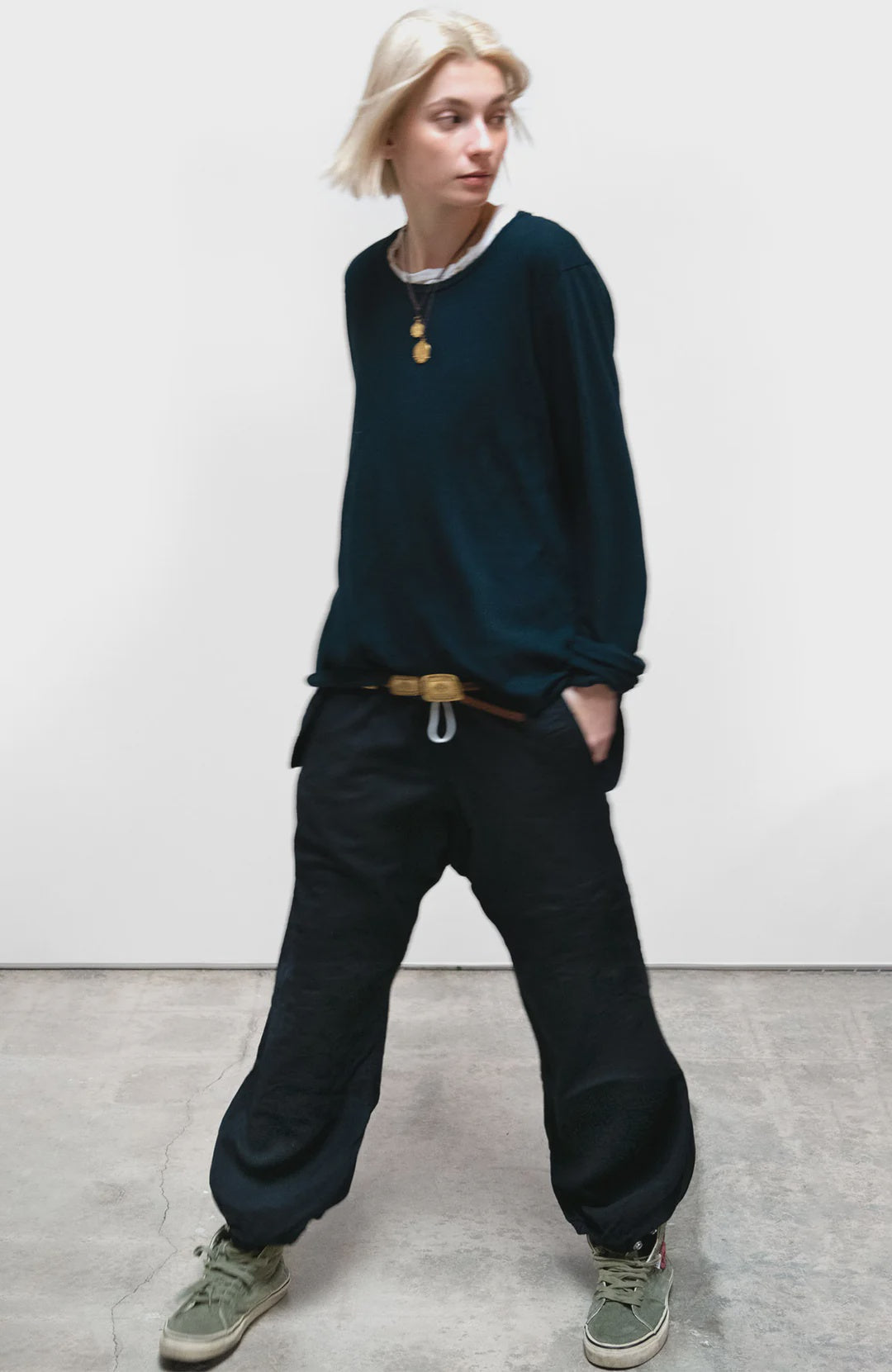 A person with short blonde hair, in a loose dark long-sleeve shirt, Free City Cashmere/Wool Flap/Snap Pant (black), and green sneakers stands on a concrete floor against a plain white background.