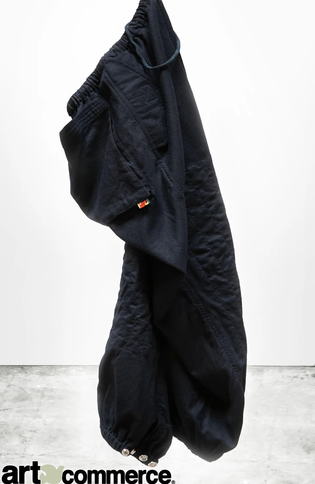 A pair of Cashmere/Wool Flap/Snap Pants by Free City (sparrow, LLC), loose-fitting and black with an elastic waistband and cuffs, hangs against a plain white background. The "art commerce" logo is at the bottom left corner.