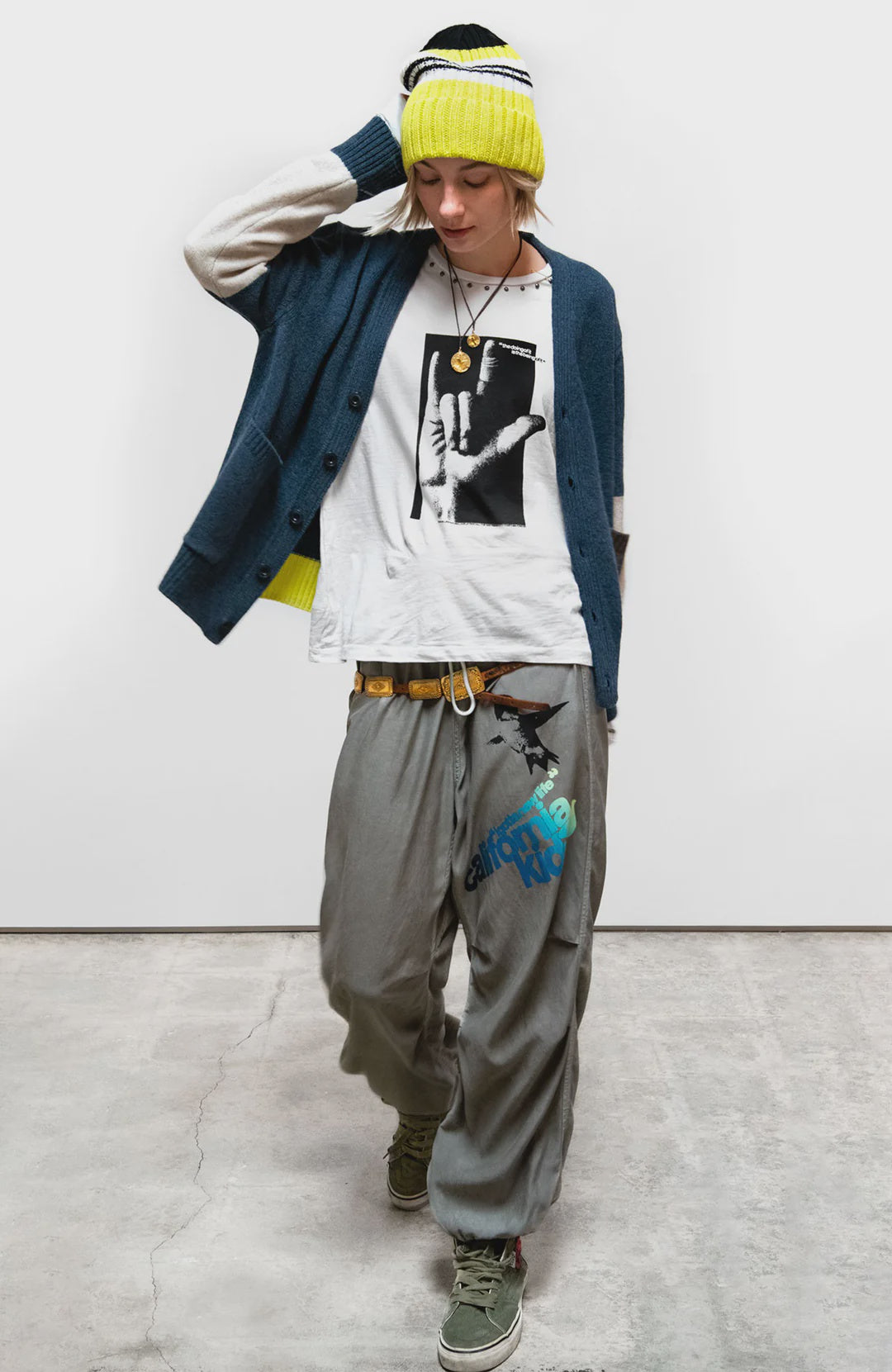 A person wearing a yellow beanie, Free City Cashmere Lux Cardigan, graphic white T-shirt, loose gray pants with blue text, and green sneakers stands indoors on a concrete floor, looking down with one hand on their head.