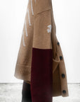 A tan and maroon Cashmere Lux Cardigan by Free City (sparrow, LLC), featuring white patterns, hangs on a hook against a white wall. "Art + commerce" appears below, evoking Loro Piana-inspired unisex elegance.
