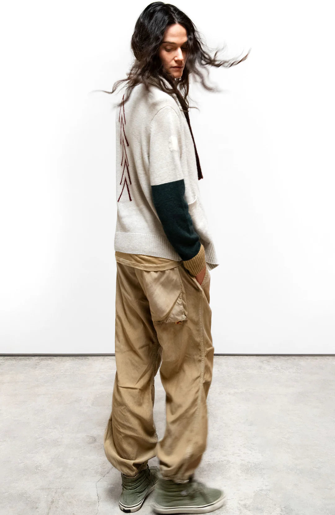 A person with long wavy hair wears a light sweater with dark sleeves, Free City Flap/Snap LG MATTE/SATIN AirJump Pant in custom sunfade wash, and green sneakers, standing in profile against a plain white wall, looking down.