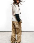 A person with long wavy hair wears a light sweater with dark sleeves, Free City Flap/Snap LG MATTE/SATIN AirJump Pant in custom sunfade wash, and green sneakers, standing in profile against a plain white wall, looking down.