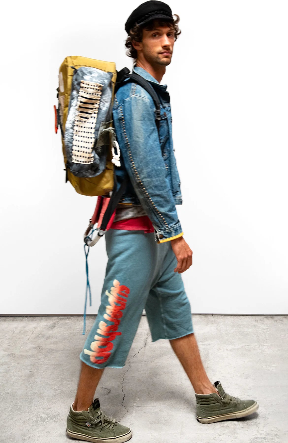 A man wearing a denim jacket, Free City (sparrow, LLC) Sparrow Homestud Yumm Cutoffs with red and white graphics, green sneakers, a black cap, and a large handmade backpack walks across a concrete floor against a plain white background.