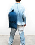 A person in loose indigo clothing and green shoes faces away, carrying a large blue Free City Indigo Sherpa Blanket/Indigo Blues bag with black text and wearing a black beret, set against a plain white background.