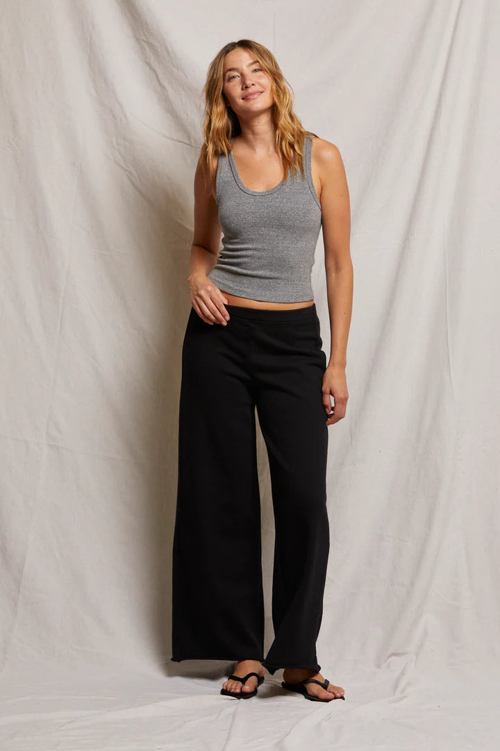 A woman in Perfectwhitetee's gray cotton blend tank top and Amber Wide Leg Terry pants stands against a neutral backdrop. She wears beige sandals, has wavy hair, and gently smiles with her hands at her sides.