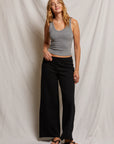 A woman in Perfectwhitetee's gray cotton blend tank top and Amber Wide Leg Terry pants stands against a neutral backdrop. She wears beige sandals, has wavy hair, and gently smiles with her hands at her sides.