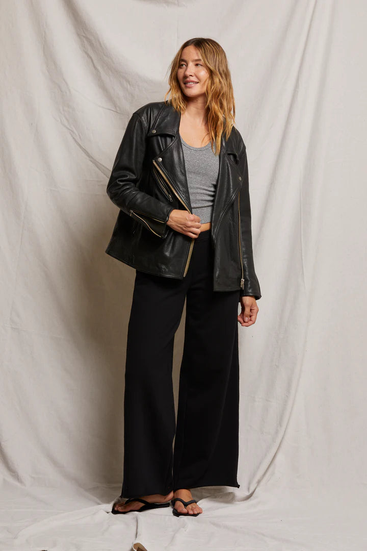 A woman stands smiling in a black leather jacket over a gray top, chic Amber Wide Leg Terry pants from Perfectwhitetee, and flip-flops. She poses gracefully against a light draped fabric backdrop.