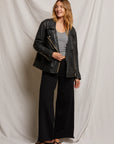 A woman stands smiling in a black leather jacket over a gray top, chic Amber Wide Leg Terry pants from Perfectwhitetee, and flip-flops. She poses gracefully against a light draped fabric backdrop.