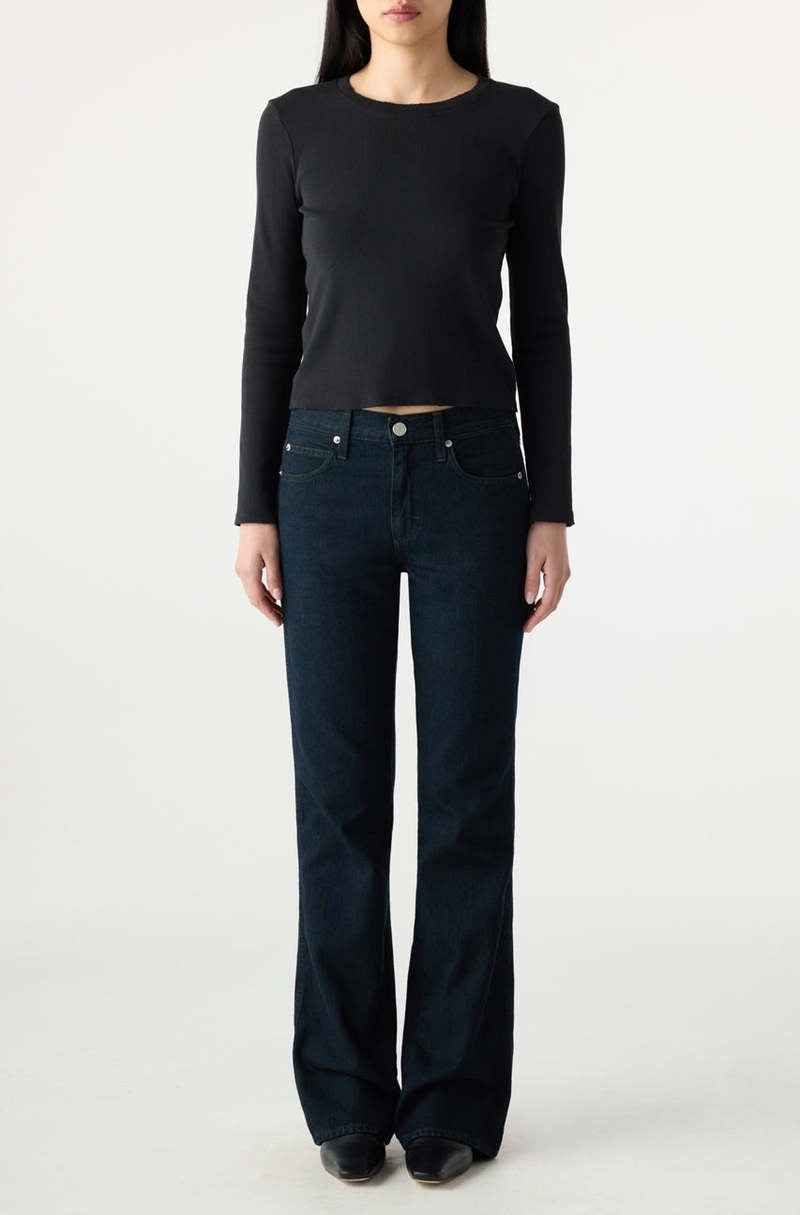 A person stands facing forward, wearing a black long-sleeve top, AMO Denim Marcenia Boot Cut dark blue jeans, and black shoes against a plain light background.