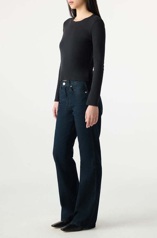 A person stands in a neutral pose wearing a fitted black long-sleeve top, black shoes, and dark blue AMO Denim Marcenia Boot Cut jeans against a plain light background. Their face is not fully visible.