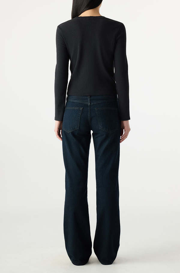 A person with long dark hair, seen from behind, wears a black long-sleeve top and AMO Denim's Marcenia Boot Cut jeans, standing against a plain light background.