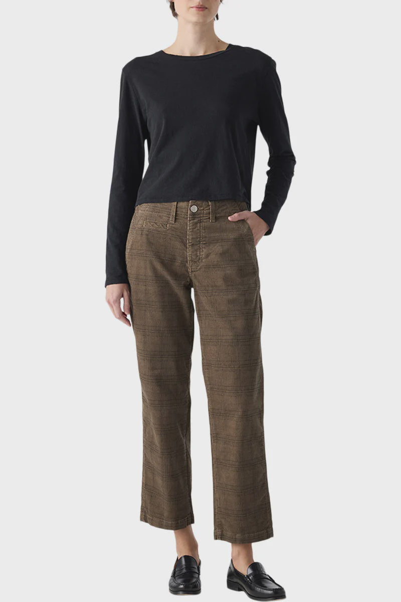 Wearing a black long-sleeve top, AMO Denim's Lillian Plaid Cord wide-leg pants, and black loafers, a person stands against a plain light background with one hand in their pocket.