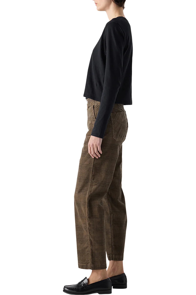 A person stands in profile against a white background wearing the AMO Denim Lillian Plaid Cord pants with a long-sleeve black shirt and black loafers.