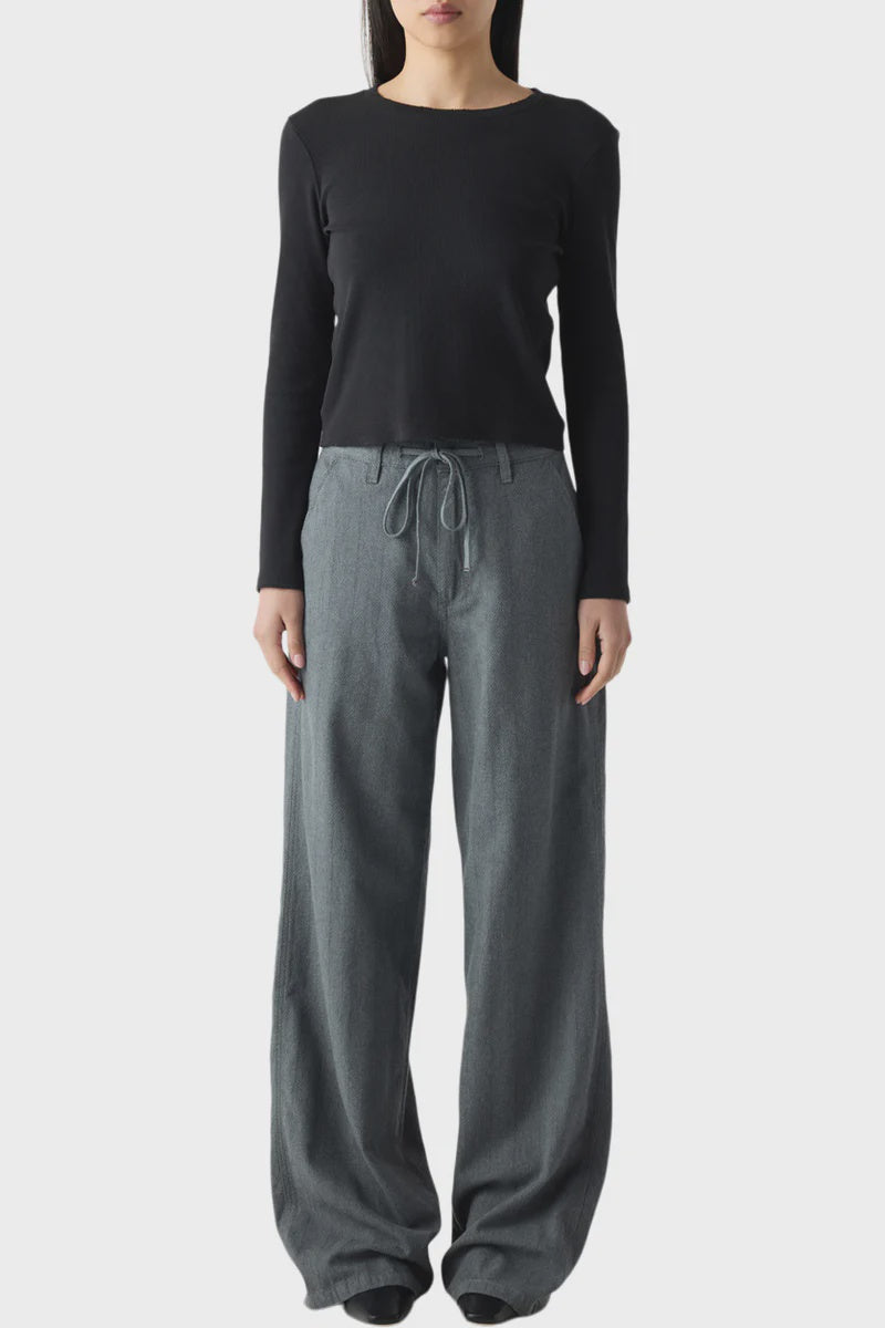 A woman stands facing forward, wearing the AMO Denim Nora Pant—loose-fitting, high-waisted drawstring pants—with a fitted black long-sleeve top. Her straight dark hair is down and her arms rest at her sides.