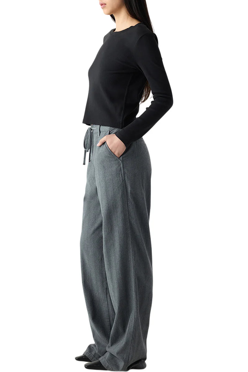 A woman stands in profile against a white background, wearing the AMO Denim Nora Pant—loose-fitting gray cotton pants—with a black long-sleeve top and her hands in her pockets.
