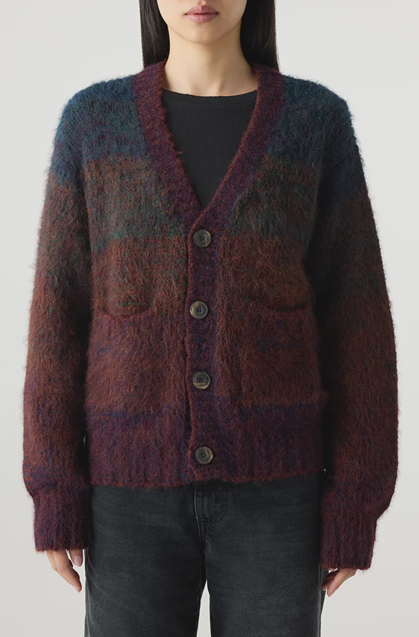 A person stands against a plain light background wearing the AMO Denim Nancy Sweater, a relaxed fit V-neck ombré cardigan in purple and burgundy over a black top, paired with dark jeans.