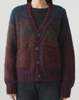 A person stands against a plain light background wearing the AMO Denim Nancy Sweater, a relaxed fit V-neck ombré cardigan in purple and burgundy over a black top, paired with dark jeans.